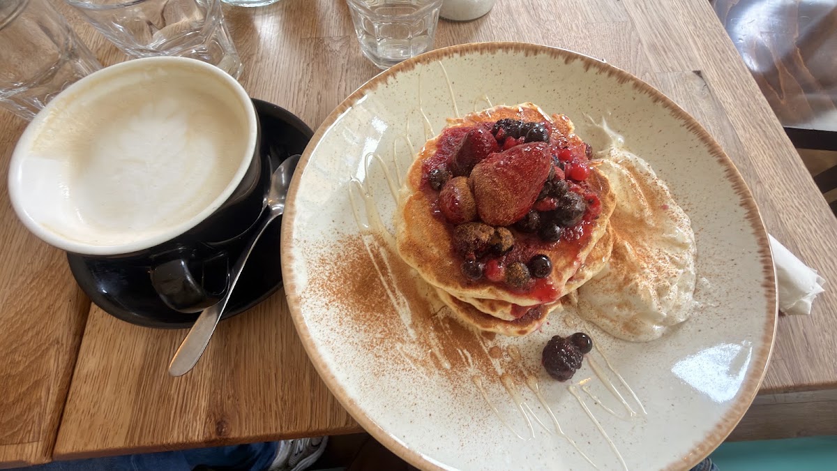 A pancake stack with berry compote, honey drizzle, cinnamon and cream, beside a flat white.