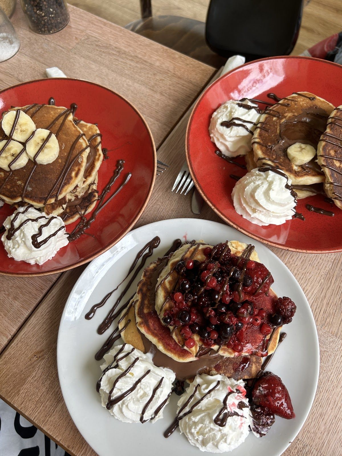 Three plates of pancakes: banana and chocolate on red plates, berries and chocolate on white.