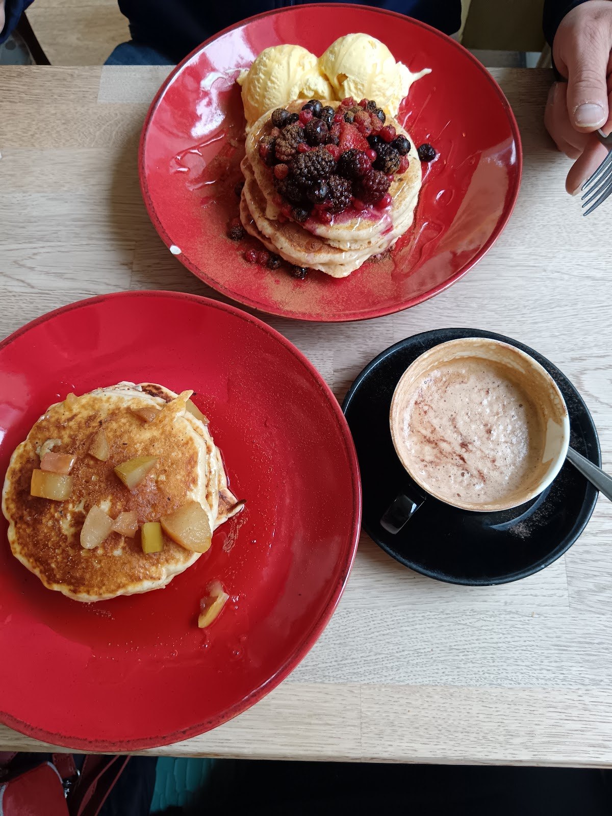 Pancakes with caramelised apple on a red plate, beside a cinnamon-dusted latte.